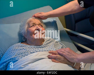 Femme âgée de vieillesse contenu souriant confortable dans le lit d'hôpital de soins à la maison avec la main réconfortante de l'infirmière de soignant Banque D'Images
