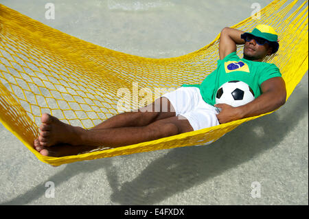 Joueur de football brésilien portant drapeau brésilien hat relaxing in Hammock Beach avec un ballon de foot Banque D'Images