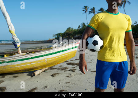 Joueur de football brésilien au Brésil couleurs holding soccer ball en face du bateau de pêche sur la plage du Brésil Nordeste Banque D'Images