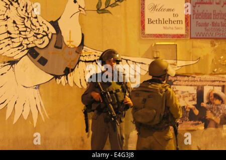 Bethléem, CISJORDANIE PALESTINE- 2014/07/09- Deux soldats israéliens attendre derrière un mur, en tant que jeunes Palestiniens jeter des pierres. Derrière les soldats, l'infâme et toujours anonymes du célèbre artiste Bansky graffiti de l''Dove de la paix" est derrière eux. Pour les huit nuits de suite, d'intenses affrontements ont éclaté à Bethléem près du Tombeau de Rachel, également connu sous le nom de Checkpoint 300. En cette nuit, de jeunes Palestiniens ont lancé des pierres d'une autre rue qui mène à la tour de garde militaire. La violence n'a pas cessé depuis la semaine dernière de l'enterrement de Muhammad Abu Khdeir et il n'y a aucun signe qu'il va mourir dans les prochaines semaines Banque D'Images