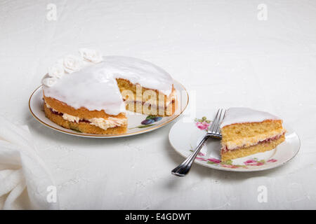 Gâteau éponge victoria iced in blanc avec des roses à la main, sur une assiette, une tranche de gâteau sur une plaque à côté(44a de 44) Banque D'Images