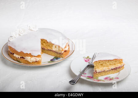 Gâteau éponge victoria iced in blanc avec des roses à la main, sur une assiette, une tranche de gâteau sur une plaque à côté de 44(35) Banque D'Images