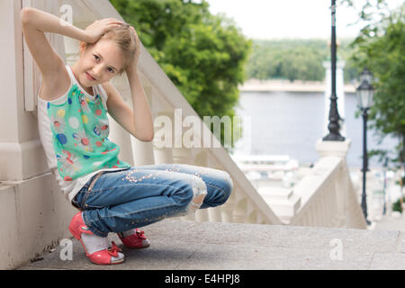 Jolie petite fille espiègle jouant dehors accroupi avec ses mains sur sa tête sur un atterrissage dans un vol d'escalier extérieur donnant sur une voie navigable souriant à la caméra. Banque D'Images