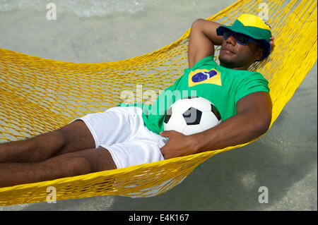 Joueur de football brésilien portant drapeau brésilien hat relaxing in Hammock Beach avec un ballon de foot Banque D'Images