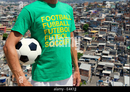 Homme debout dans brésilien de football international t-shirt holding soccer ball en face de taudis favela de Rio de Janeiro d'arrière-plan Banque D'Images