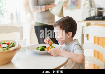 Boy (6-7) Bien manger le dîner, la mère en arrière-plan Banque D'Images