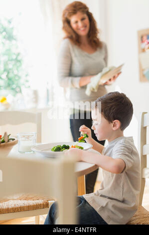 Boy (6-7) Bien manger le dîner, la mère en arrière-plan Banque D'Images