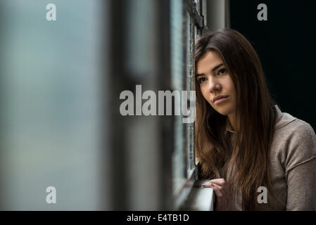 Portrait of young woman standing in front of fenêtre, rêver et à la recherche dans la distance, Allemagne Banque D'Images