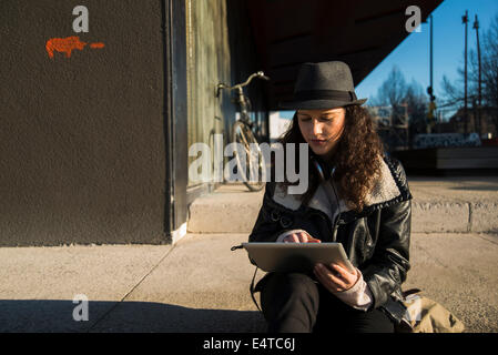 Teenage girl sitting on curb de trottoir à l'extérieur, le port de Fedora et using tablet computer, Allemagne Banque D'Images