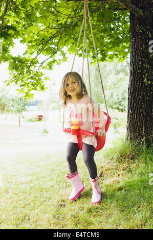 3 ans, fille en bottes de caoutchouc assis dans red swing en cour arrière, looking at camera and smiling, Suède Banque D'Images