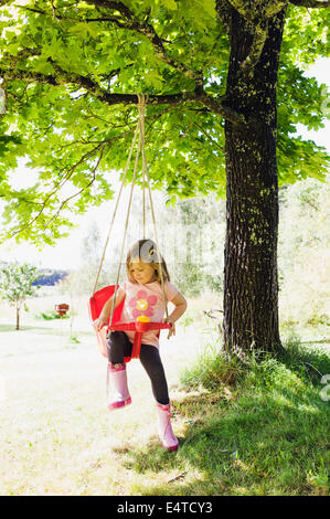 3 ans, fille en bottes de caoutchouc assis dans balançoire rouge en arrière-cour, la Suède Banque D'Images