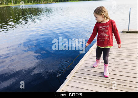 3 ans, fille en chemise rouge sur un quai tenant un bâton et jouant dans l'eau, la Suède Banque D'Images