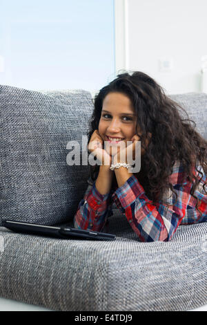 Close-up portrait of teenage girl at home, lying on sofa with tablet computer, looking at camera and smiling, Allemagne Banque D'Images
