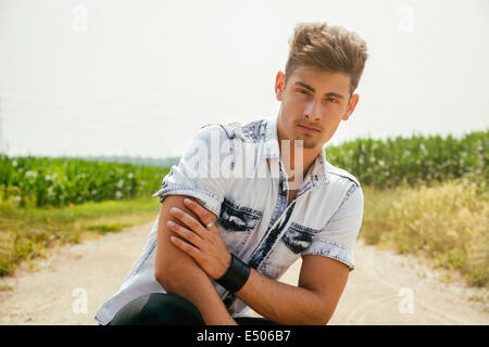 Beau jeune homme assis au milieu de campagne chemin de terre, looking at camera Banque D'Images