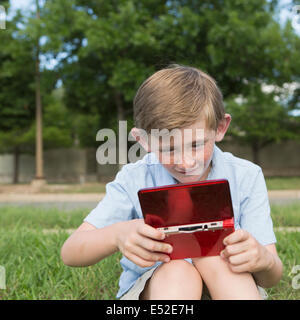 Un jeune garçon assis sur l'herbe de la lecture d'un jeu électronique de poche. Banque D'Images