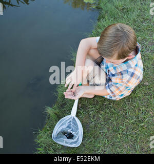 Un jeune garçon assis sur une rive du fleuve, l'examen d'un petit poisson, un tiddler il retirés dans un filet. Banque D'Images