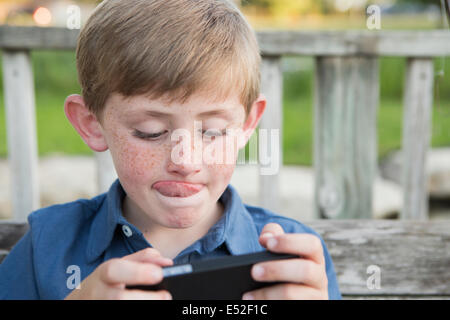 Un jeune garçon à l'aide d'une tablette électronique ou un jeu et se concentrer, s'en tenir sa langue. Banque D'Images