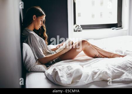 Vue latérale du beautiful young woman reading book sur le lit chez lui. Modèle féminin en lingerie, assis sur le lit de lire un roman. Banque D'Images