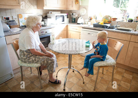 Grand-mère assise à la table avec son petit-fils (de 3 ans) alors qu'il boit une tasse de lait et mange un biscuit, Royaume-Uni Banque D'Images