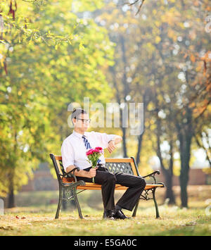 Jeune homme tenant des fleurs et du contrôle du temps assis sur un banc dans le parc Banque D'Images