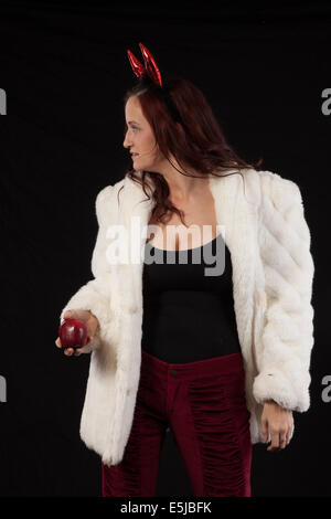 Woman in devil horns et veste de fourrure blanche, noire et rouge avec chemisier Pantalon souriant et tenant une pomme dans la tentation, Banque D'Images