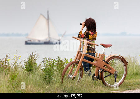 Pingjum Pays-bas, Rameau, Vélo. Le Dutch Design vélo en bois. Woman looking at bateau à voile avec des jumelles Banque D'Images