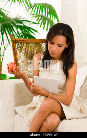 Belle jeune femme élégante assise dans living using digital tablet et manger une pomme Banque D'Images