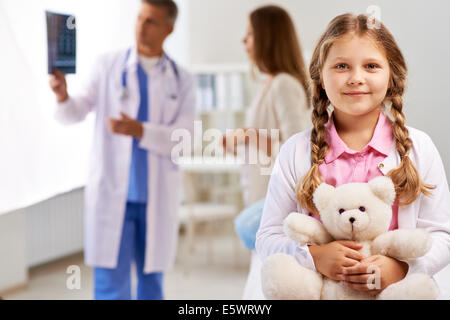 Happy girl with teddy bear looking at camera sur fond de médecin montrant les résultats radiologiques à sa mère Banque D'Images