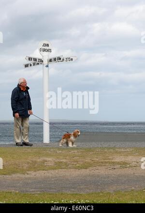Un homme promène son chien passé la balise marquant la fin de la masse terrestre écossais, John O' Groats, Caithness, Highlands, Scotland Banque D'Images