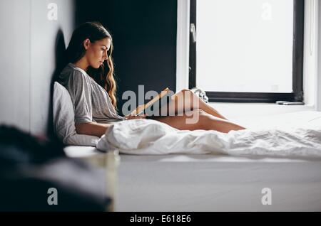 Vue latérale d'une grenaille belle jeune femme assise sur son lit la lecture d'un roman intéressant. Modèle des femmes de race blanche dans la chambre. Banque D'Images