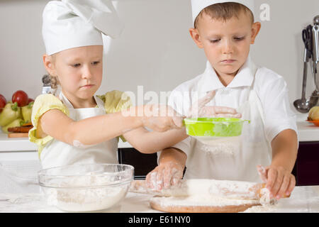 Petit garçon et fille en blanc toques et tabliers la cuisson dans la cuisine faire la pâte et travailler en équipe comme la jeune fille se répand la farine comme le garçon de rouleaux sur la pâte. Banque D'Images