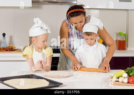 Mère enseigne la manière de faire la pizza pour les enfants portant des vêtements de chefs à la cuisine Banque D'Images