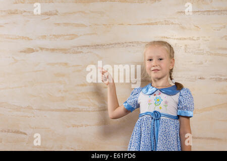 Cute blonde girl porte une robe avec motif floral wile pointant vers le mur derrière elle en bois Banque D'Images