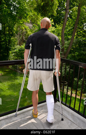Homme blessé avec une jambe dans le plâtre et des béquilles se remettre de blessures sur un balcon donnant sur une forêt Banque D'Images