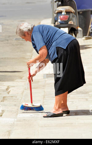 Scène de rue vieille femme âgée citoyenne se pliant pour balayer le trottoir à l'extérieur de sa maison à Polignano a Mare ville dans la province de Bari Apulia Puglia Italie eu Banque D'Images