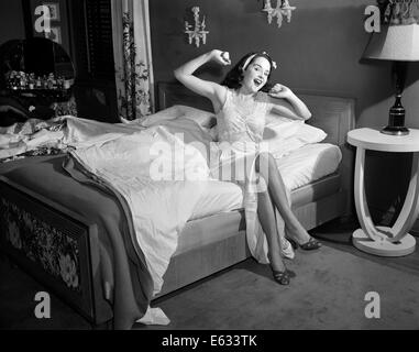 Années 1950, JEUNE FEMME EN Robe de nuit CHAUSSONS ASSIS EDGE BED LOOKING AT CAMERA ÉTIREMENT BÂILLEMENT se réveiller Banque D'Images