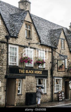 Femme marche passé le cheval noir public house sous la pluie, du centre-ville, Cirencester, Cotswolds, Gloucestershire, Angleterre, Royaume-Uni. Banque D'Images