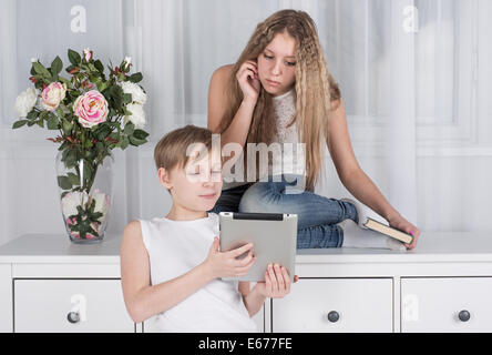 Fille assise sur kamod avec un livre et un garçon debout à côté d'elle et montre digital tablet Banque D'Images