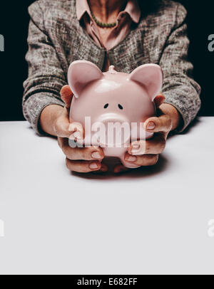 Portrait studio shot of senior female mains tenant un coin bank tout en étant assis à une table. Vieille Femme tenant une tirelire. Banque D'Images