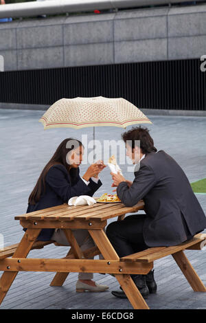 Young couple sitting on bench eating lunch s'abritant sous le parapluie dans la pluie à Southbank, Londres en Août Banque D'Images