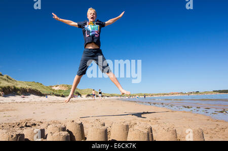 Un garçon sautant au dessus d'un château de sable sur la plage de Bamburgh, Northumberland, Angleterre. Banque D'Images