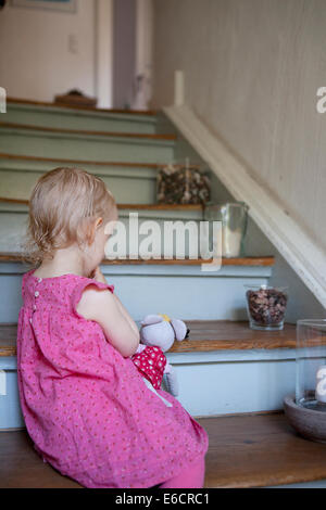 Jeune fille dans une robe rose, assis sur les escaliers dans une maison avec son doudou. Banque D'Images