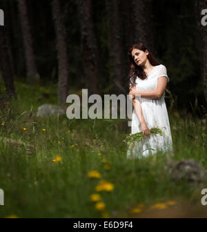 Femme aux cheveux longs en robe blanche et white hat dans la main marcher en forêt tenant un bouquet de fleurs Banque D'Images