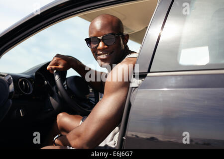 Portrait of happy young guy dans sa voiture à la caméra en souriant. Modèle masculin africain portant des lunettes de soleil. L'homme musclé en voiture. Banque D'Images