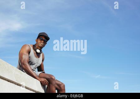 Jeune homme africain élégant assis contre le ciel bleu. Homme portant des lunettes de soleil et chapeau avec un casque à la recherche à l'appareil photo. Banque D'Images