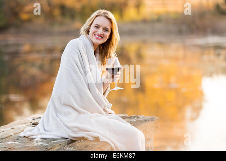 Smiling young woman sitting on pier et de boire le vin rouge au coucher du soleil Banque D'Images