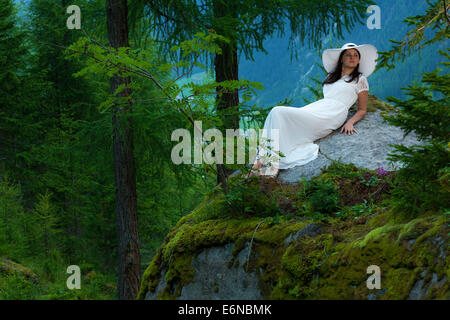 Caucasian woman in white dress with white hat posé sur une grosse pierre dans la forêt Banque D'Images