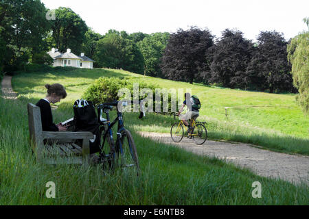 Des promenades en vélo cycliste passé une femme lisant un livre assis sur un banc en été sur Hampstead Heath North London NW3 UK KATHY DEWITT Banque D'Images