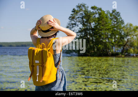 Une jeune femme avec un sac à dos jaune tenant son chapeau devant un lac et arbres en été Banque D'Images