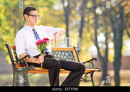 Jeune homme tenant des fleurs et du contrôle du temps assis sur un banc dans le parc Banque D'Images